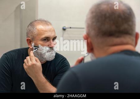 Uomo maturo dai capelli grigi che restyling la sua barba in casa utilizzando il rasoio Foto Stock