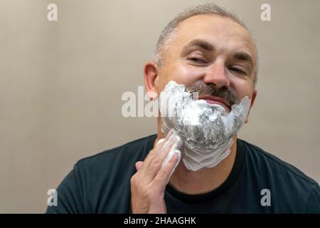 Ritratto di bel bianco-capelli beared uomo di mezza età applicando schiuma da barba per rifinire la barba Foto Stock