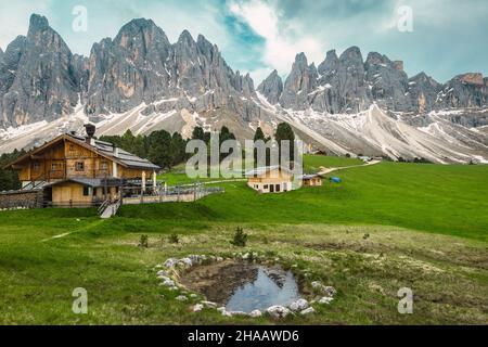 Maestoso paesaggio alpino primaverile, case in legno sui campi verdi e laghetto con montagne sullo sfondo, gruppo di Geisler - Odle, Alto A. Foto Stock