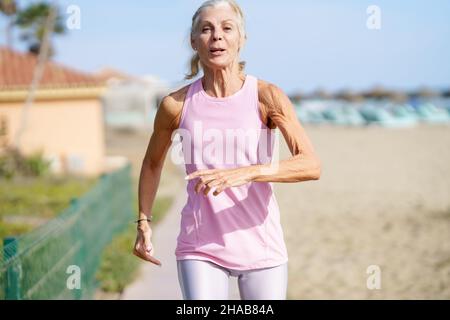 Donna matura che corre lungo la riva della spiaggia. Donna più anziana che fa sport per mantenere la misura Foto Stock