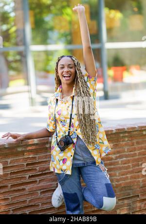 Donna nera con trecce africane, sollevando il braccio in gioia. Ragazza che tiene una macchina fotografica. Foto Stock