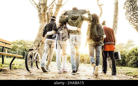 Gruppo di amici urbani che camminano nel parco di pattinare della città con retroilluminazione al tramonto - concetto di gioventù e amicizia con i giovani multirazziali che si divertono Foto Stock