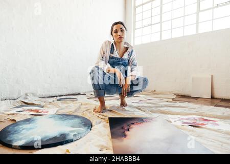 Una pittrice di talento femminile che si schiava nel bel mezzo dei suoi dipinti. Giovane artista fiducioso che guarda la fotocamera nella sua galleria d'arte. Donna giovane creativa Foto Stock