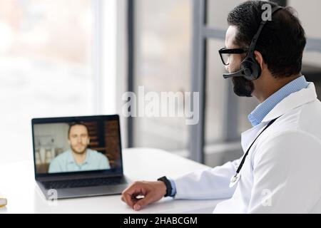 Appuntamento medico sulla distanza. Il medico indiano maschio che usa il laptop per l'incontro in linea con il paziente, ascolta i reclami, discute il programma di assicurazione con l'uomo giovane Foto Stock