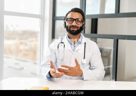 Fidato MD maschile indiano indossare cuffie, occhiali e camice medico dando consultazione online, tenendo videochiamata, il medico guarda la fotocamera e parlare a distanza. Telemedicine, vista webcam Foto Stock