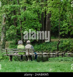 Una coppia anziana siede su una panca di legno da un piccolo ruscello in un parco circondato da verde estivo. Foto Stock