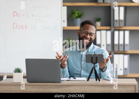 Felice giovane africano americano barbuto ragazzo insegnante in occhiali lavora al portatile e guarda la webcam Foto Stock