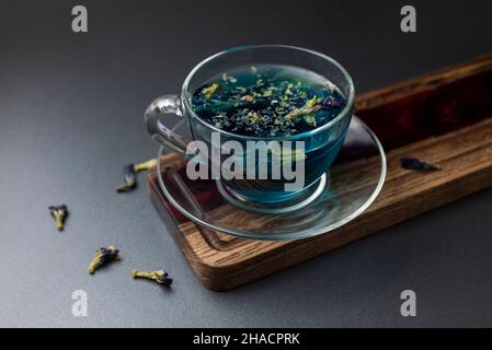 Tè blu tailandese Anchan in una tazza di vetro trasparente con foglie di tè. Bevanda aromatica calda dall'Asia su un asse di legno con fiori secchi Foto Stock