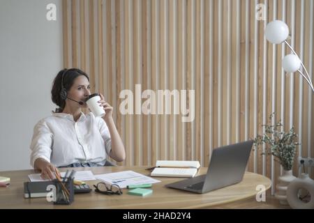 Soddisfatto positivo spagnolo imprenditore femminile in cuffia wireless bere caffè in ufficio Foto Stock