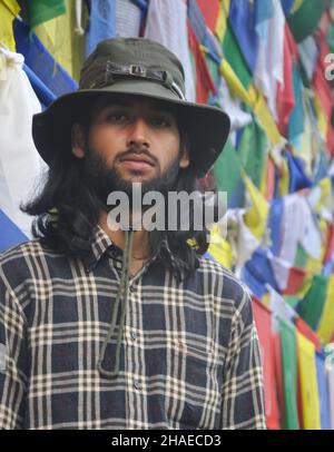 Un turista maschio barbuto e dai capelli lunghi che guarda la macchina fotografica mentre si alza contro le bandiere di preghiera buddiste Foto Stock