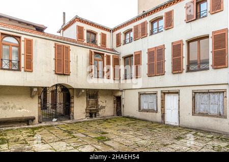 Un cancello in ferro battuto e una vecchia pompa dell'acqua in pietra si trovano in un cortile deserto nel centro storico di Saint Chamond, visti attraverso un portale a volta, dipartimento della Loira, Francia. Il muschio copre i ciottoli del cortile. Foto Stock