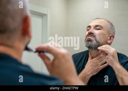 Uomo maturo dai capelli grigi che restyling la sua barba in casa utilizzando il rasoio Foto Stock