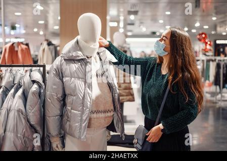 Giovane donna in maschera di faccia sceglie abiti caldi nel grande magazzino, in piedi vicino manichino vestito in puffy giacca invernale e maglione, l'acquisto di nuovo vestito stagionale. Shopping durante covid19 pandemic Foto Stock