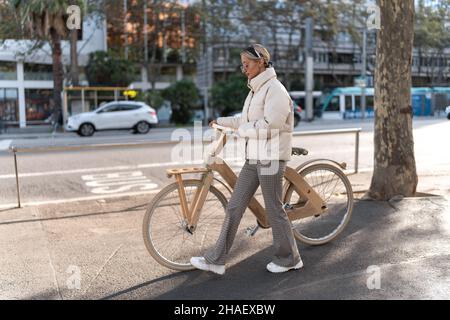 Donna a corpo pieno in outerwear che spinge la bicicletta eco-compatibile mentre cammina sul marciapiede vicino alla strada nella giornata di sole in città. Foto Stock