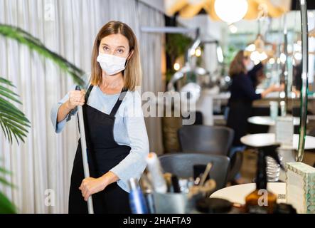 Giovane donna che indossa grembiule e maschera protettiva viso spazzando pavimento in barbiere. Compiti quotidiani di parrucchiere. Precauzioni necessarie durante coronaviru Foto Stock