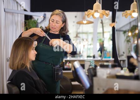 Donna anziana focalizzata parrucchiere taglio capelli di femmina cliente con forbici in moderno parrucchiere. Foto Stock