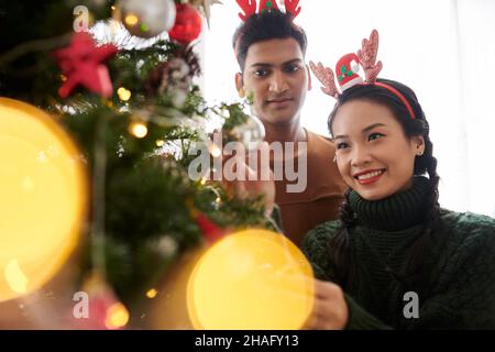 Felice giovane coppia variegata che decora albero di Natale per la celebrazione Foto Stock