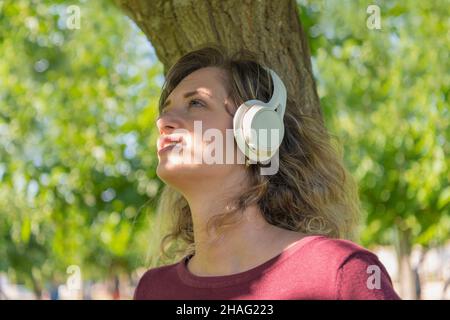 Donna ascolta la musica con le cuffie wireless mentre si appoggia su un albero in un parco Foto Stock