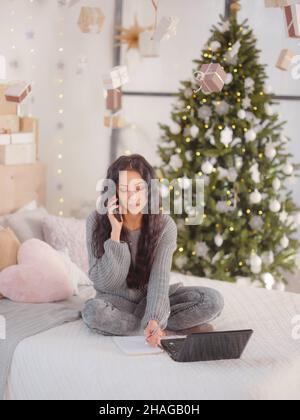 Giovane bella donna caucasica con capelli scuri in morbido maglione a maglia grigio usando il laptop sul letto in camera con albero di Natale. Decide le questioni relative alle prossime vacanze, ordini di consegna, effettua acquisti Foto Stock