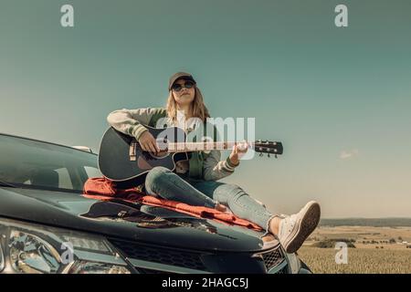 una giovane ragazza che si cimenta con la chitarra sul cofano della sua auto durante il viaggio in campagna Foto Stock