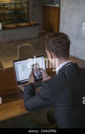 Il giovane uomo d'affari effettua videochiamate sul cellulare e lavora con il laptop in un bar. Bell'uomo caucasico in abito formale che lavora in un caffè Foto Stock