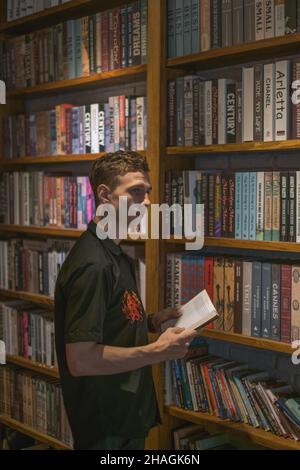 Giovane uomo caucasico in piedi vicino alle mensole di legno con libri nel caffè. Tenere e leggere il libro. Stile vintage in una caffetteria. Libreria. Foto Stock