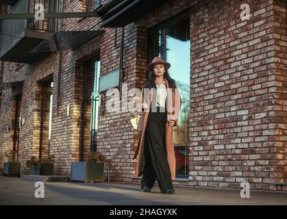 Donna asiatica d'affari in un lungo cappotto beige e cappello cammina lungo la strada in uno stile loft, sullo sfondo di un muro di mattoni, stile retrò Foto Stock