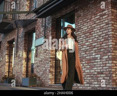 Donna asiatica d'affari in un lungo cappotto beige e cappello cammina lungo la strada in uno stile loft, sullo sfondo di un muro di mattoni, stile retrò Foto Stock