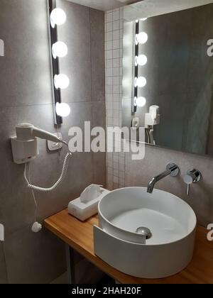 Il bagno dispone di un lavandino, un asciugacapelli appeso alla parete accanto, una fila di luci sulla parete e uno specchio Foto Stock