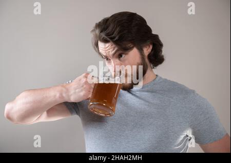 l'uomo bearded beve birra con una tazza grande su uno sfondo grigio Foto Stock