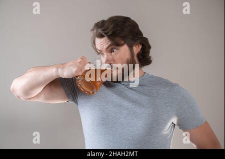 l'uomo bearded beve birra con una tazza grande su uno sfondo grigio Foto Stock