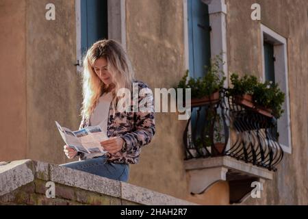 Una ragazza bionda siede su un parapetto di pietra vicino ad un vecchio edificio sullo sfondo di un balcone vintage in fiori e legge una rivista Foto Stock