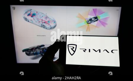 Persona che tiene il telefono cellulare con il logo del produttore di auto croato Rimac automobili sullo schermo di fronte alla pagina web. Mettere a fuoco sul display del telefono. Foto Stock