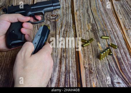 Le cartucce traumatiche sono sparse su uno sfondo di legno marrone. Le mani tengono una clip a pistola e una pistola Foto Stock