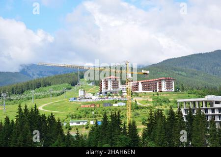 Una gru a torre costruisce una stazione sciistica in montagna in estate, sullo sfondo di montagne, foreste e alberghi in costruzione. Gli impianti di risalita Foto Stock