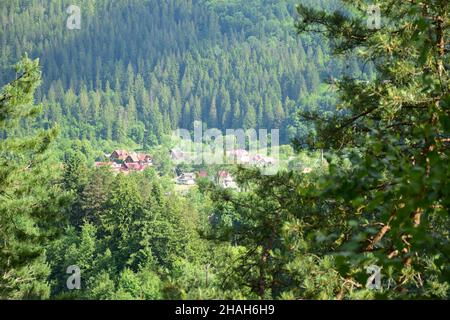 Molto più in basso, sotto la montagna, si trova un piccolo villaggio circondato da una foresta verde estiva. Fotografato dal lato attraverso il fogliame, con Foto Stock