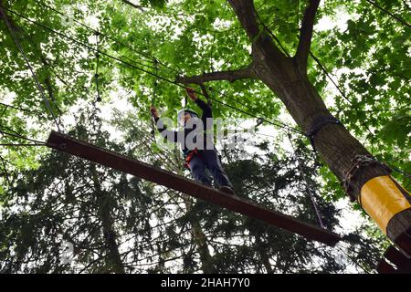 Un ragazzo adolescente in un'imbracatura di sicurezza attraversa un ponte sospeso in un parco estivo con funi Foto Stock