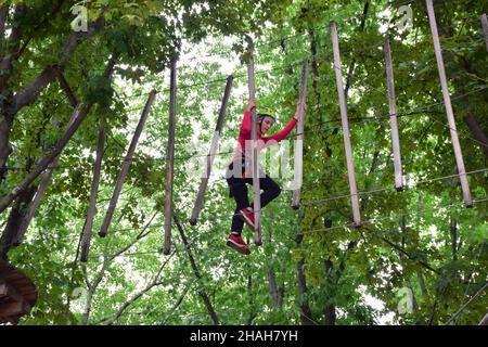 Un ragazzo adolescente in un'imbracatura di sicurezza attraversa un ponte sospeso in un parco estivo con funi Foto Stock