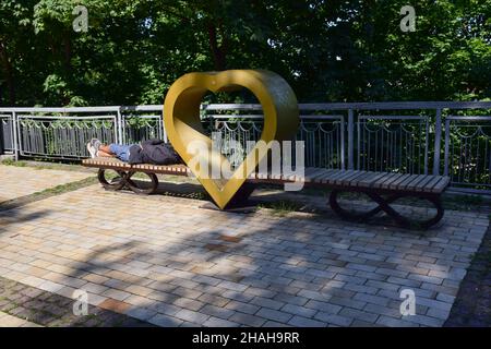 In un parco estivo, un uomo non identificato giace e riposa su una panchina sotto forma di cuore Foto Stock