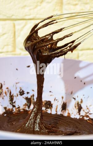 Fare il cioccolato a casa. Liquido e scuro su sfondo sfocato montato con una frusta in una piastra di plastica Foto Stock