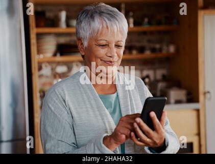 Sorridente multi-culturale anziana femmina scorrimento Internet su smartphone mentre si trova in cucina moderna. Foto Stock