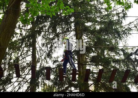 Un ragazzo adolescente in un casco di sicurezza sale una scala sospesa in un parco di divertimento in corda Foto Stock