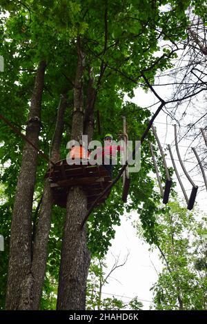 Un ragazzo adolescente in un casco di sicurezza sale una scala sospesa in un parco di divertimento in corda Foto Stock