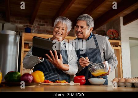 Coppia multiculturale anziana sorridente, usando il tablet per cercare ricetta in cucina moderna. Felice coppia che cucinano pasto sano. Foto Stock