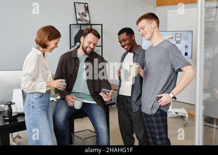 Giovane uomo d'affari che mostra qualcosa sul telefono cellulare ai suoi colleghi mentre bevono caffè in ufficio Foto Stock
