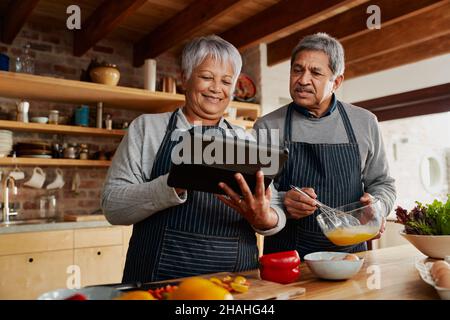 Coppia multiculturale anziana sorridente, usando il tablet per cercare ricetta in cucina moderna. Lifestyle felice cucina pasto sano. Foto Stock