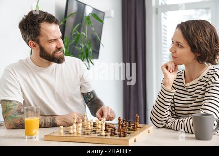 Felice giovane coppia bianca che gioca a scacchi seduti al tavolo a casa parlando Foto Stock