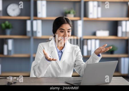 Sorridente giovane medico indiano femminile nei gesti di cappotto bianco guardare il laptop webcam e comunica con il paziente Foto Stock