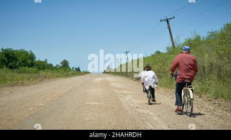 Vista posteriore del nonno con i nipoti in bicicletta che ritornano dalla scuola sulla strada sterrata Foto Stock