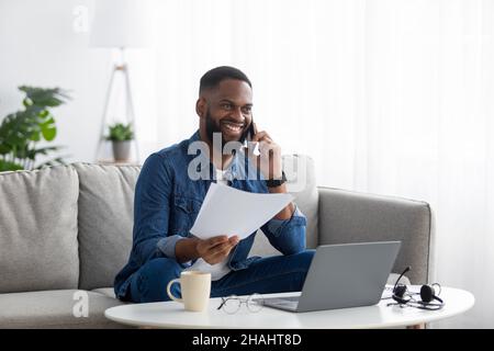 Felice attraente millenario africano americano barbuto ragazzo lavora con documenti e computer, chiamate per telefono Foto Stock